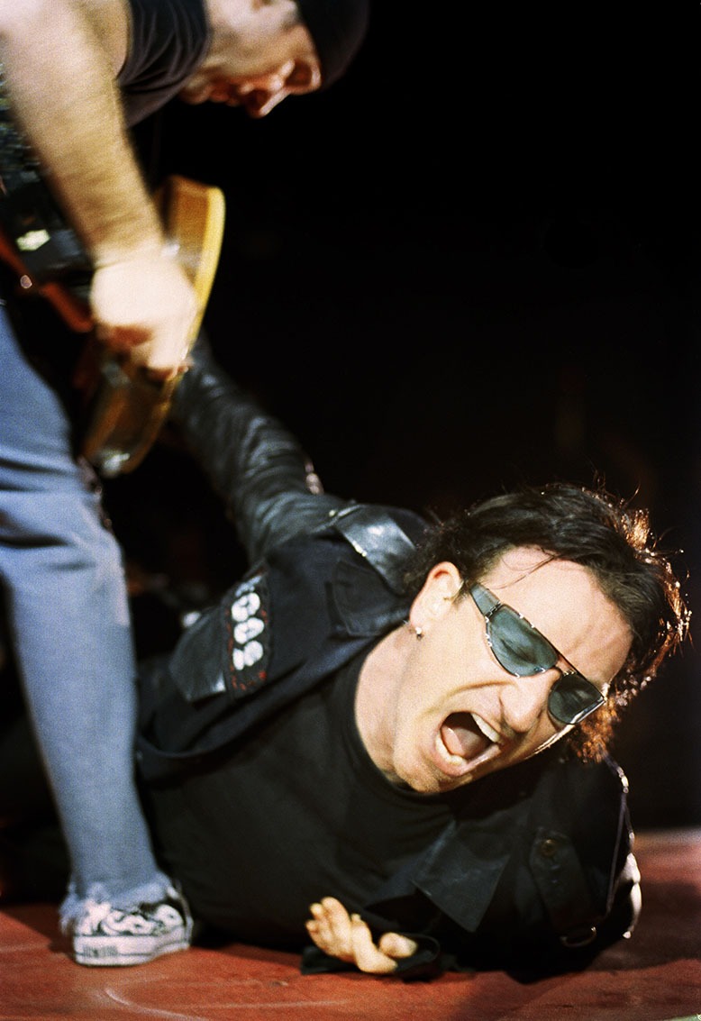Photographer Jay Blakesberg stands above Bono of U2, who is lying on the ground of a stage during a November 2021 performance