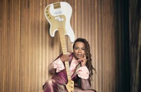 Divinity Roxx, sitting with one hand on her face, wields an upside-down guitar in the air with the other hand