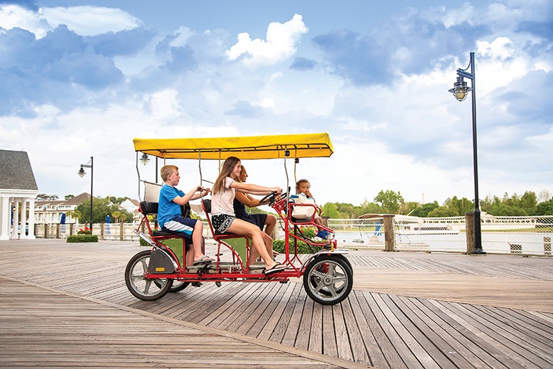 A family rides a surrey bicycle on a boardwalk.