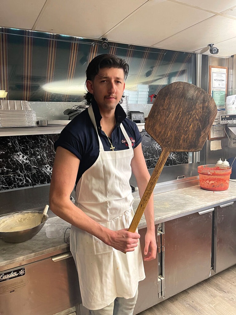 Salvatore Mandreucci dons an apron and holds a wooden pizza paddle in the kitchen