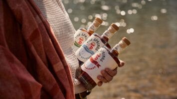 Woman holds three bottles of Five Springs Bourbon in front of a lake sparking with sunlight