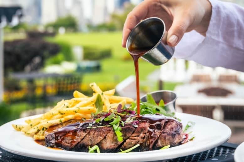 Steak sauce being poured onto a plate of steak and fries. The New York City skyline is visible in the background.