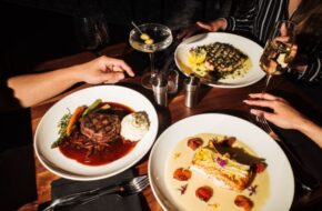 A table with cocktails and dinner plates full of food. Two hands rest on the table.