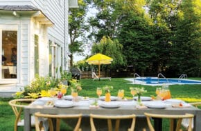 The backyard dining and lounge areas of a white Spring Lake colonial, featuring lush greenery, a pool, bright yellow umbrellas, and a table ready for entertaining