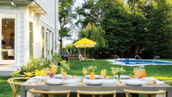 The backyard dining and lounge areas of a white Spring Lake colonial, featuring lush greenery, a pool, bright yellow umbrellas, and a table ready for entertaining