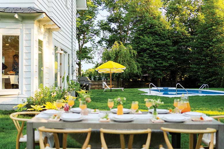 The backyard dining and lounge areas of a white Spring Lake colonial, featuring lush greenery, a pool, bright yellow umbrellas, and a table ready for entertaining