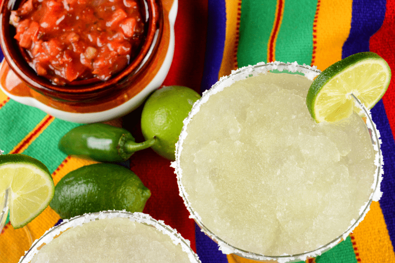 Margaritas with salt and lime on colorful tabletop with bowl of salsa