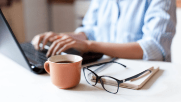 Woman typing at computer at desk with a notebook, glasses and cup of coffee