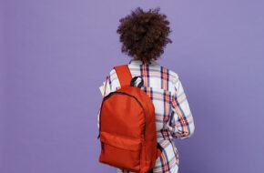 Back rear view of young girl woman of African American ethnicity wearing backpack isolated on pastel purple color background