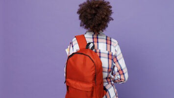Back rear view of young girl woman of African American ethnicity wearing backpack isolated on pastel purple color background