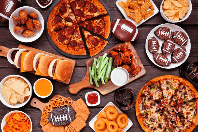 Food for Super Bowl on table including pizza, hamburgers, wings, snacks and sides
