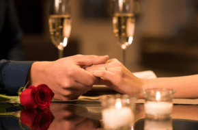 Couple holding hands at dinner table with candles, champagne glasses and rose