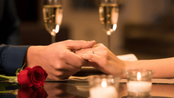 Couple holding hands at dinner table with candles, champagne glasses and rose