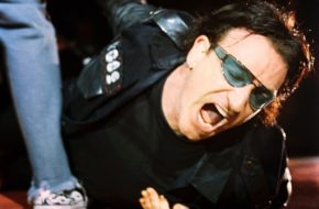 Photographer Jay Blakesberg stands above Bono of U2, who is lying on the ground of a stage during a November 2021 performance