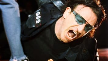 Photographer Jay Blakesberg stands above Bono of U2, who is lying on the ground of a stage during a November 2021 performance