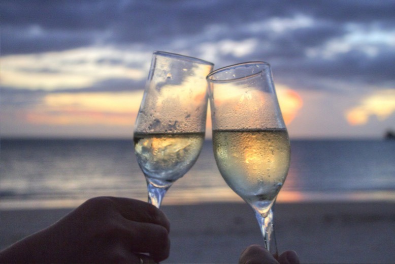Clinking glasses filled with white wine against a backdrop of a beach at sunset