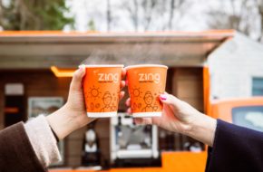 Two people toasting orange Zing coffee cups outside the orange coffee shop on wheels