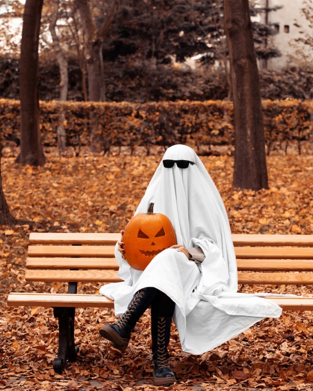 Person sitting on bench amid fall leaves, wearing a ghost costume and sunglasses and holding a jack-o'-lantern
