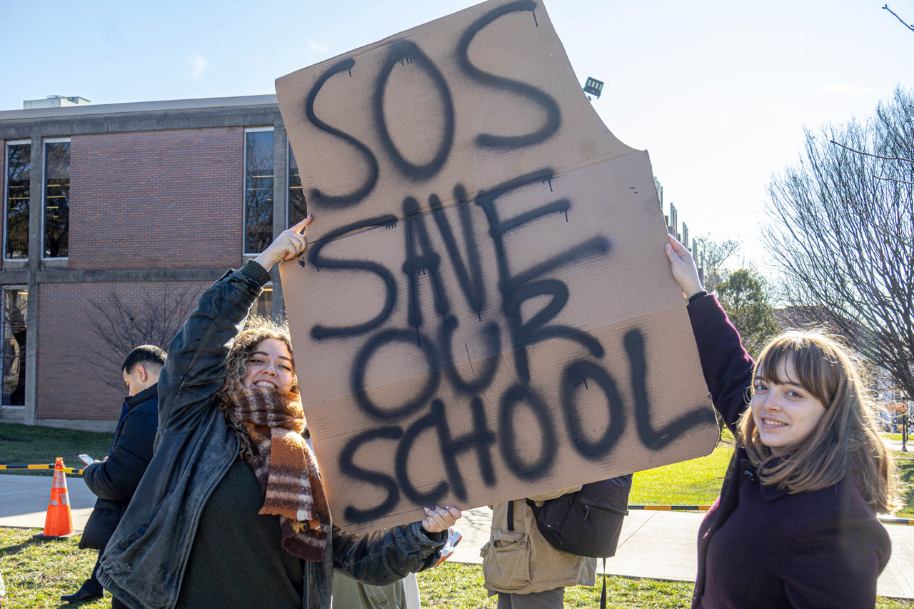 At a rally to keep humanities majors at Montclair State University, students hold a cardboard sign spray-painted in black text that reads "SOS SAVE OUR SCHOOL"