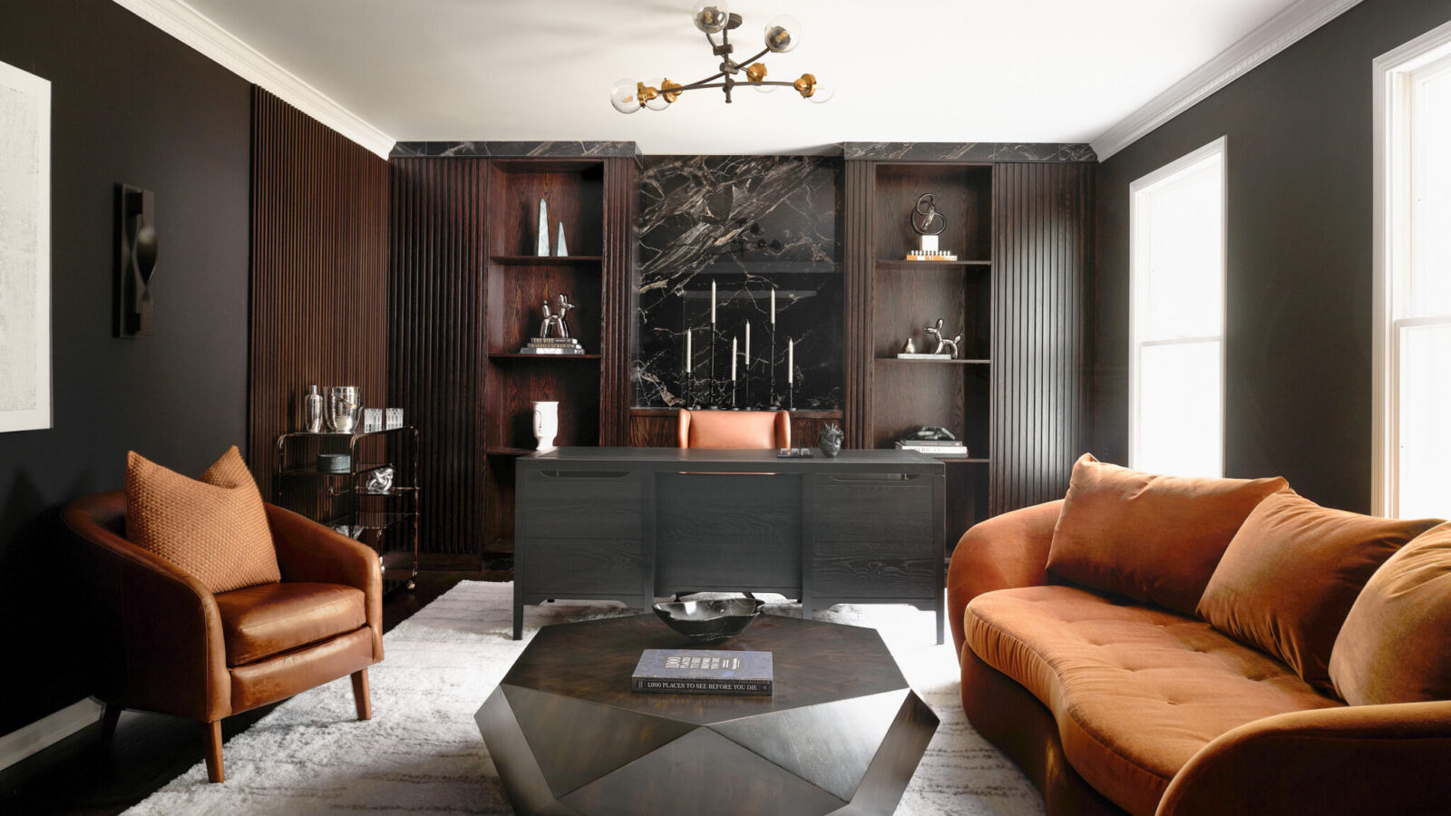 Inside a colonial home in Randolph, NJ, lush textures, sculptural lighting and organic forms soften the masculine edge of this moody home office, equally suited to work, game days and unwinding with friends. Polished Galaxy porcelain sets a dramatic backdrop for bespoke built-ins, while flanking fluted panels hide integrated doors.
