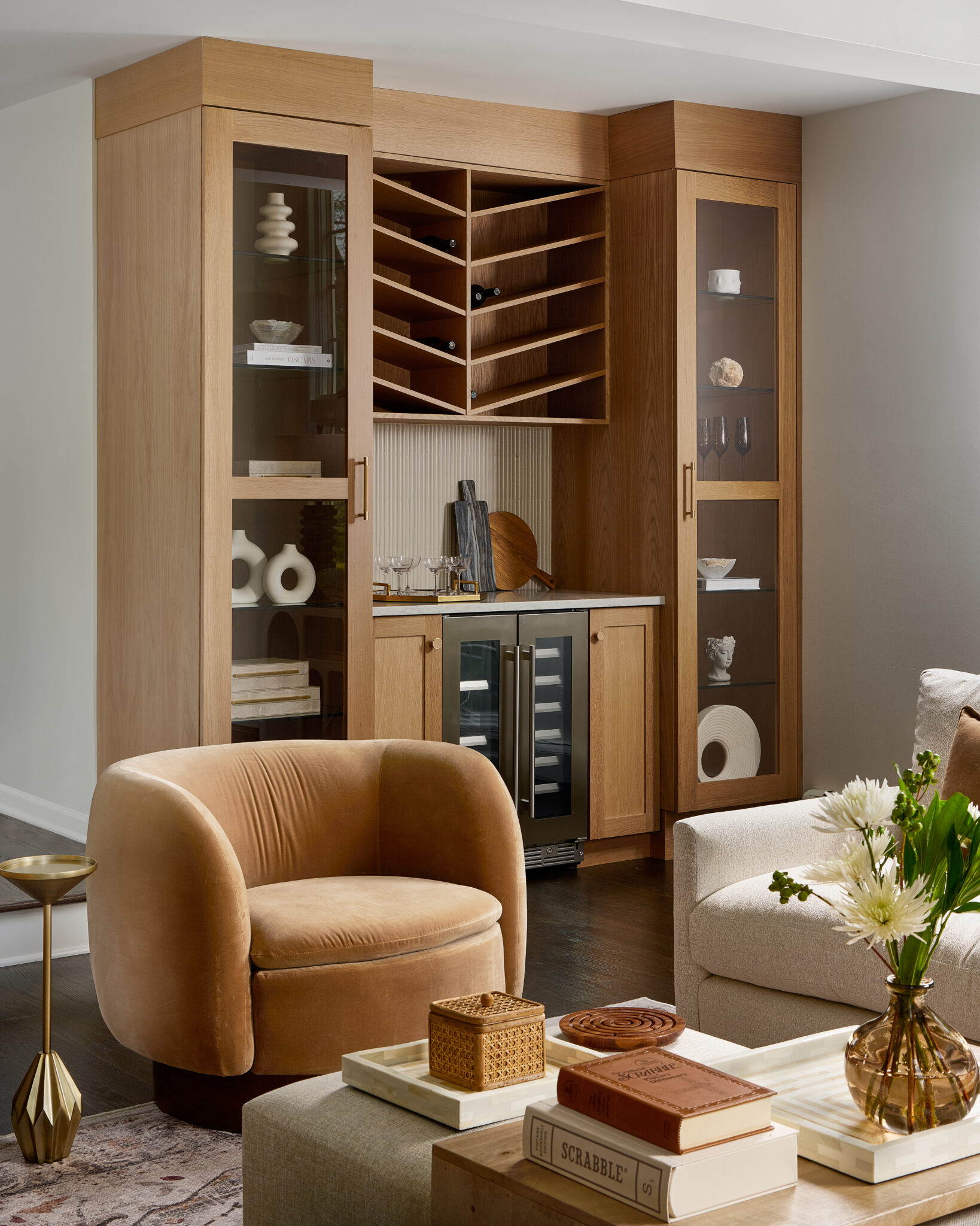 Inside a colonial home in Randolph, NJ, made-to-measure white-oak built-ins in the stepdown living room feature a beverage fridge, wine storage and glass-front cabinets to display collectibles, set against a handmade Spanish-tile backsplash. 