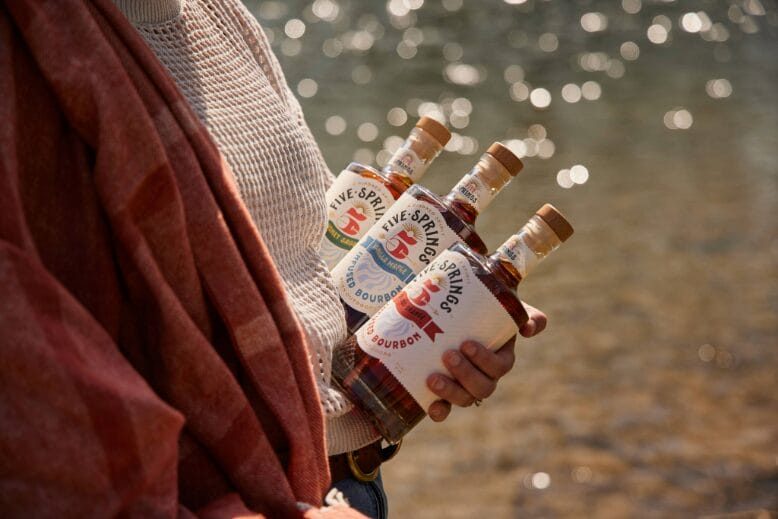 Woman holds three bottles of Five Springs Bourbon in front of a lake sparking with sunlight