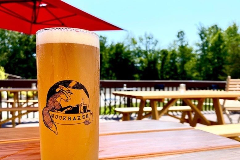 Muckraker Beermaker's Insalata ale on an outdoor table with an umbrella and trees in the background