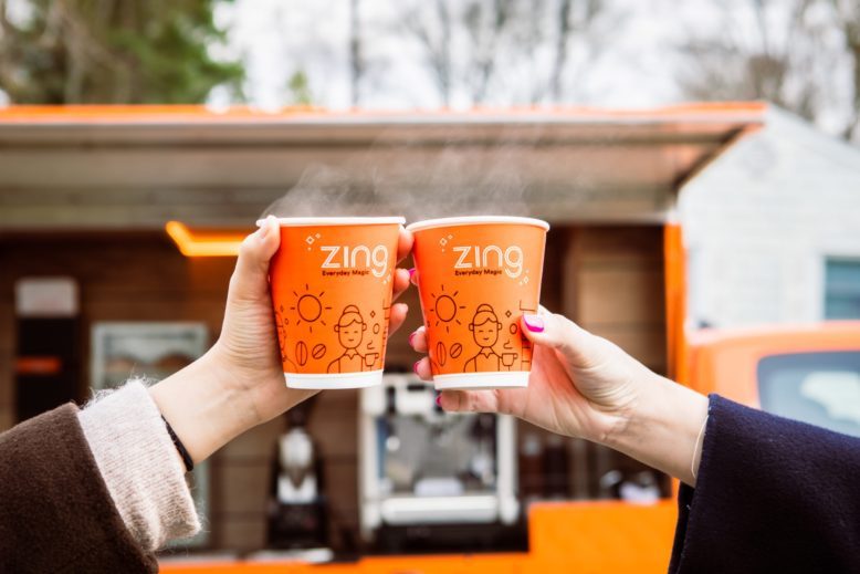 Two people toasting orange Zing coffee cups outside the orange coffee shop on wheels